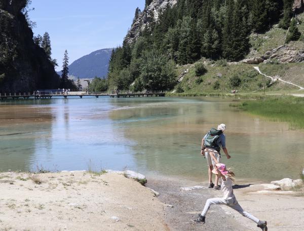 A vibrant outdoor scene shows an adult and a child by a clear, multi-hued mountain lake, with the child playfully jumping